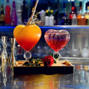 deux verres à cocktails en forme de cœur , à gauche l'un est rempli d'une boisson orange, et à droite un plus petit, est remplit d'une boisson rouge translucide, ils sont présentés sur un plateau sur un bar en inox