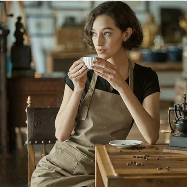 Tablier de cuisine imperméable et résistant à l'huile beige porté par une jeune fille assise dans ça cuisine buvant un thé