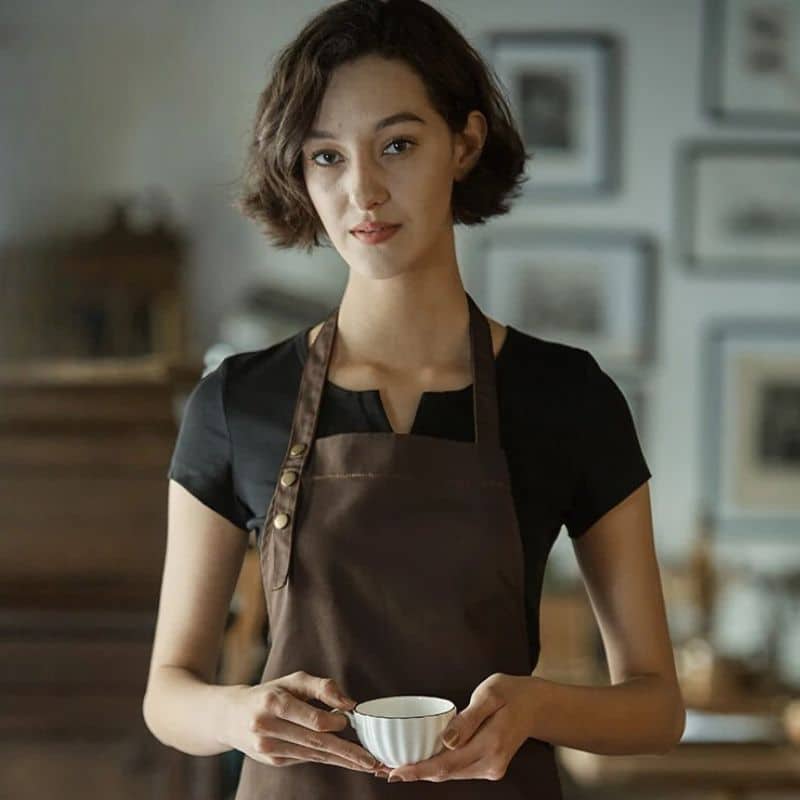 Tablier de cuisine imperméable et résistant à l'huile marron porté par une jeune fille dans ça cuisine se tenant debout et tenant une tasse de thé