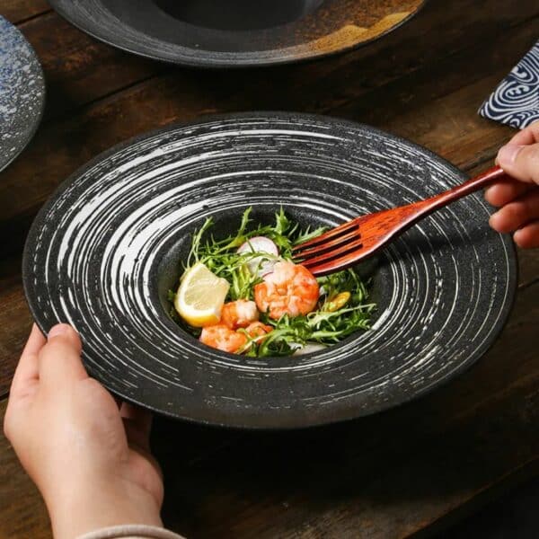 Assiette pates finition moderne, une main tiens l'assiette où il y a des aliments à l’intérieur. L'assiette est posée sur une table en bois.